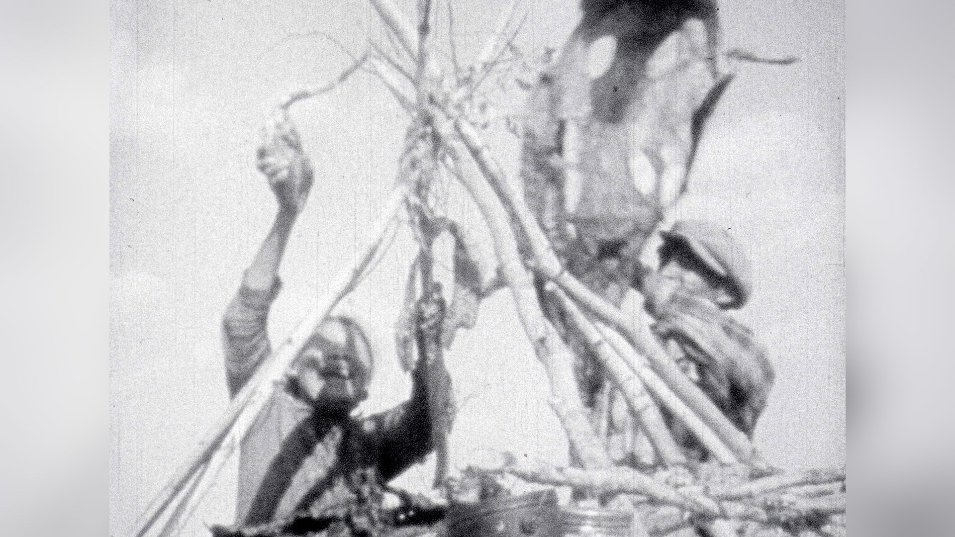 : Two figures appear assembling a structure made of wooden branches forming a point at the top. The image is a black and white film still of what appears to be archival footage.