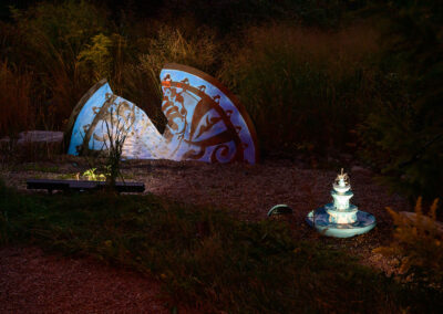A minuscule fountain and a larger-than life replica vase are lit up in a dark garden.