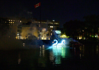 Projections of a fish and two swans emerge from a small cloud of smoke over a pond. Vari Hall is in the background.