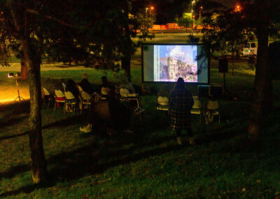 A film is projected in an outdoor cinema, with several viewers sitting on chairs.